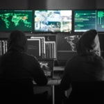 Two Hooded hackers in dark room with a bank of computer screens